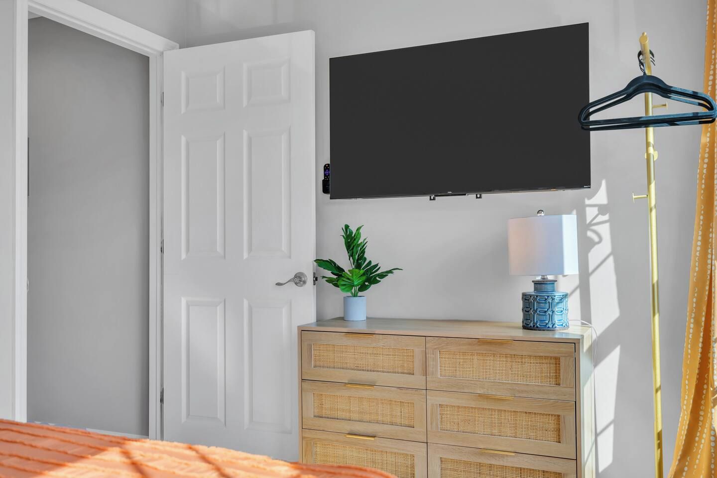 Un dormitorio con cama, televisor montado en la pared, mesita de noche con lámpara, planta en maceta y cómoda de madera.