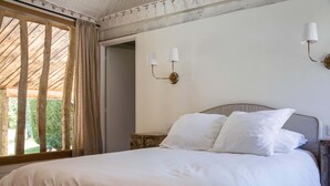 Un dormitorio con una cama grande, una mesita de noche, una lámpara de pared y vistas a la vegetación a través de una ventana.
