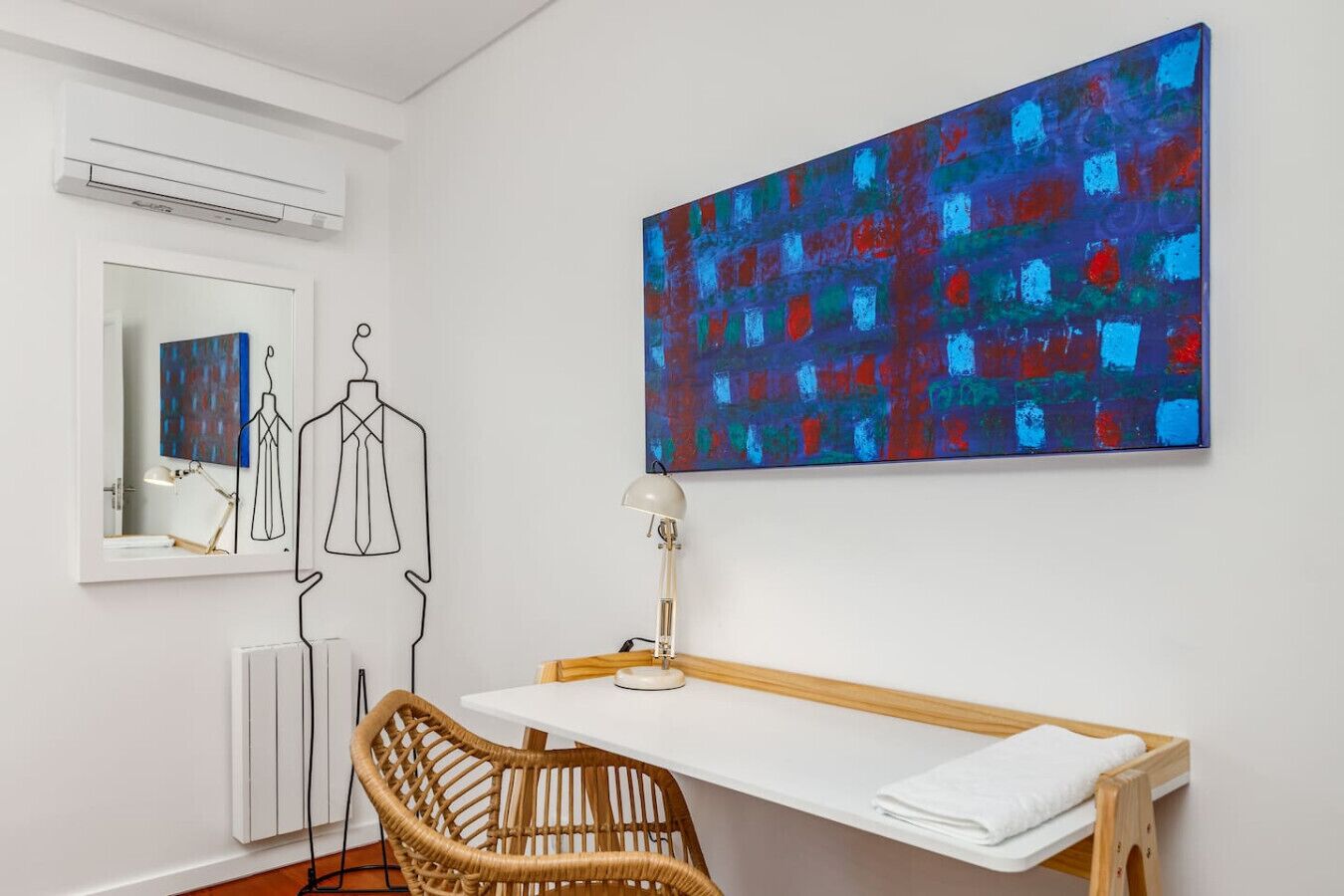 A room with a white desk, a wicker chair, a large abstract painting, a mirror, and a radiator.