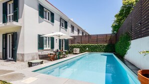 A rectangular swimming pool with a white tiled deck, a table with chairs, and a small table with a chair. - Haus Abel - with pool and garden in central Lisbon (Lisboa)