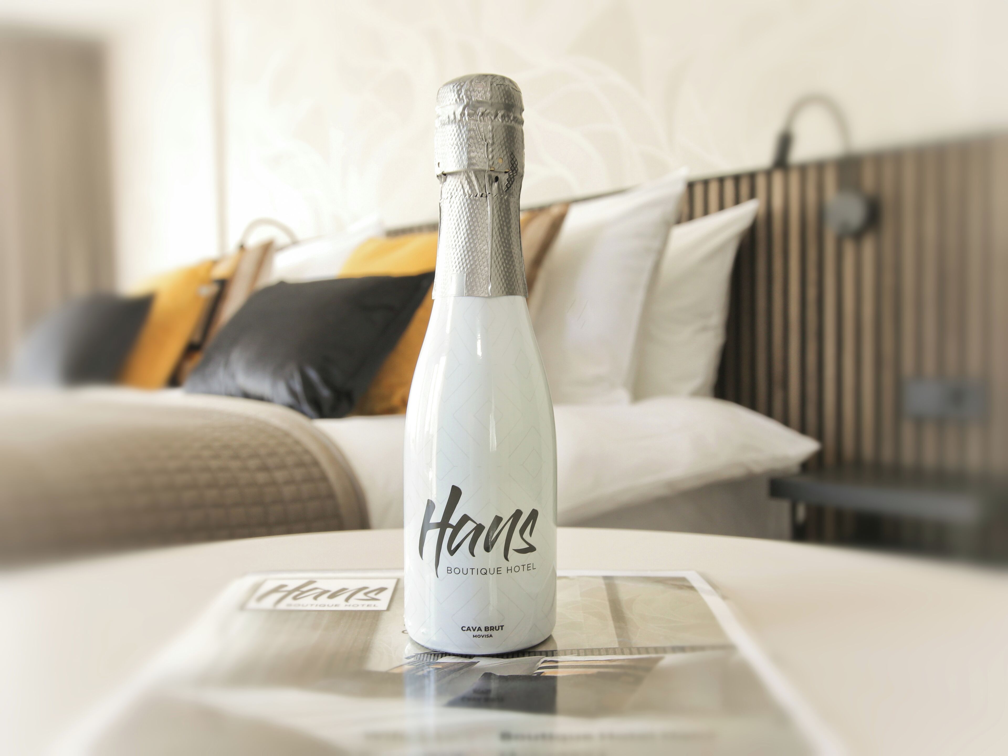 A bottle of champagne on a hotel bed with a white and gray color scheme, a bed with a black and yellow pillow, and a wooden headboard.