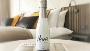 A bottle of champagne on a hotel bed with a white and gray color scheme, a bed with a black and yellow pillow, and a wooden headboard.