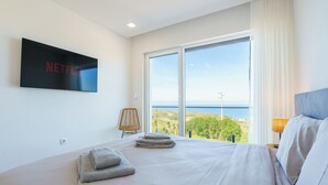 A hotel room with a large bed, a flat-screen TV mounted on the wall, a view of the sea through large windows, and a wicker chair. - Axess Villa with panoramic ocean view (Lisboa)