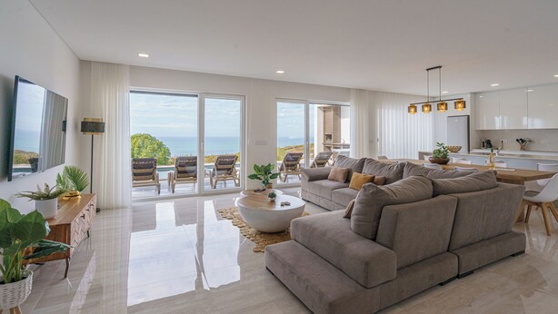 A modern living room with a large sofa, a coffee table, and a view of the ocean through large sliding doors. - Axess Villa with panoramic ocean view (Lisboa)