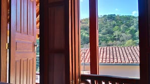 An open wooden door leading to a balcony with a view of a tiled roof and lush greenery. - Pousada Olivier da Montanha (Santo Antonio do Pinhal)