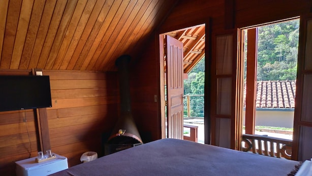 A wooden cabin room with a bed, a television, a fireplace, and a balcony with a view of trees. - Pousada Olivier da Montanha (Santo Antonio do Pinhal)