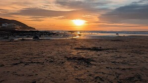 Une plage au coucher du soleil, avec une côte rocheuse et des bâtiments en arrière-plan.