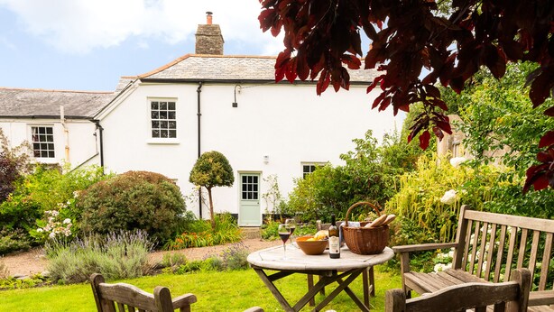 Une maison blanche avec un jardin, une table ronde entourée de chaises et une bouteille de vin.