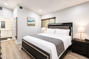 A modern bedroom with a bed, bedside tables, a nightstand, a window with blinds, and a bathroom visible through an open door.