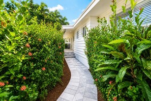 A pathway flanked by lush green hedges with red flowers, leading to a building with a white exterior and a covered entrance.