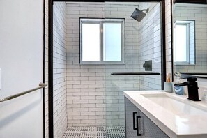 Un baño moderno con paredes de azulejos blancos, cabina de ducha de vidrio, ventana y lavamanos blanco con grifo negro.