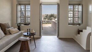 A modern living room with a sofa, a small round table, and a view through an open door to an outdoor balcony. - Frescoes Luxury Suites (Santorini)