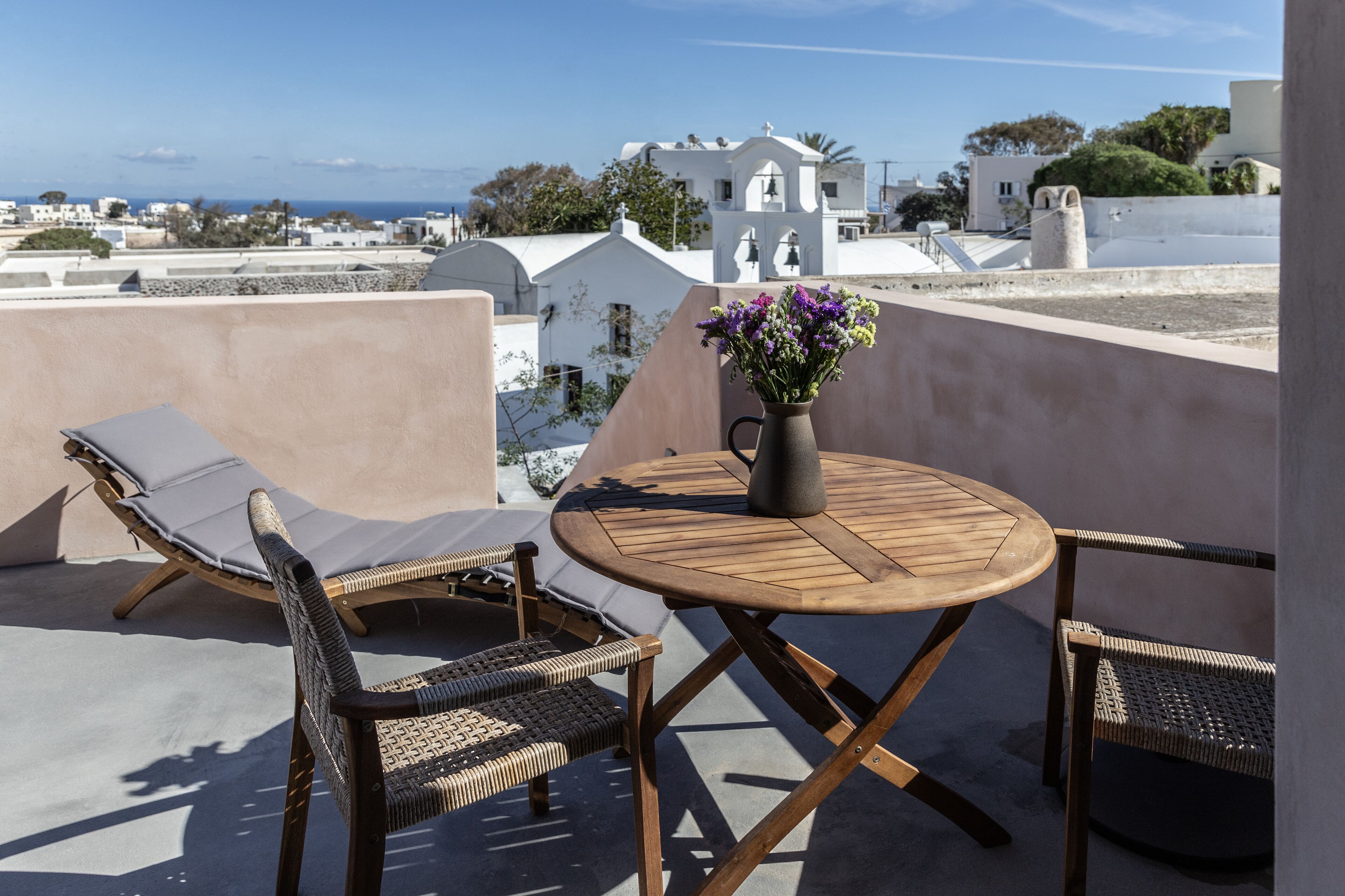 A rooftop terrace with a round wooden table, two wicker lounge chairs, and a vase with flowers.