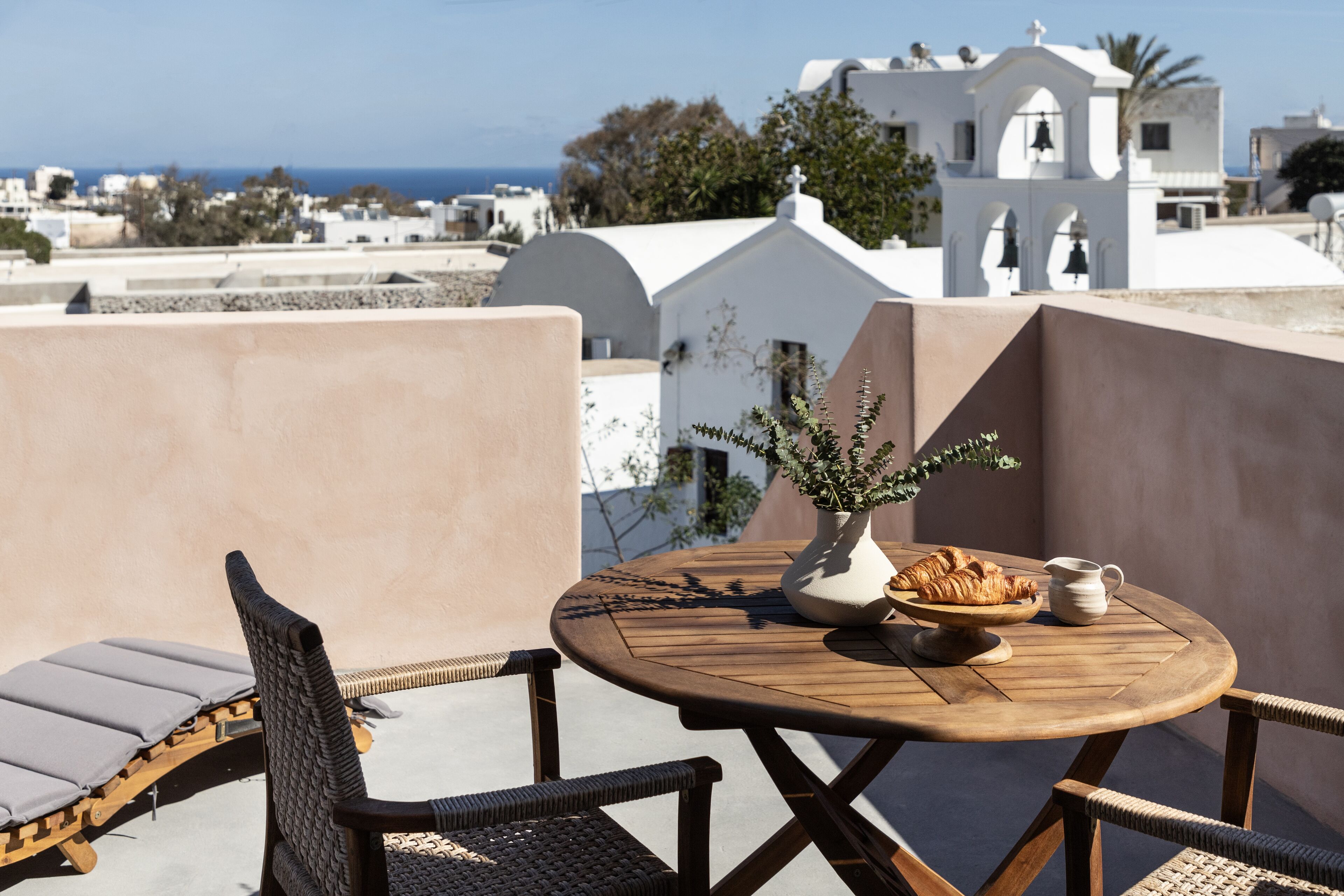 A round wooden table with a white vase, a bowl of croissants, and a white pitcher on a balcony overlooking a coastal town with white buildings and a clear sky.