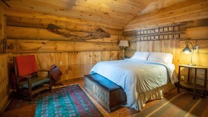A cozy wooden cabin bedroom with a bed, a chair, a bench, a lamp, and a ceiling fan.