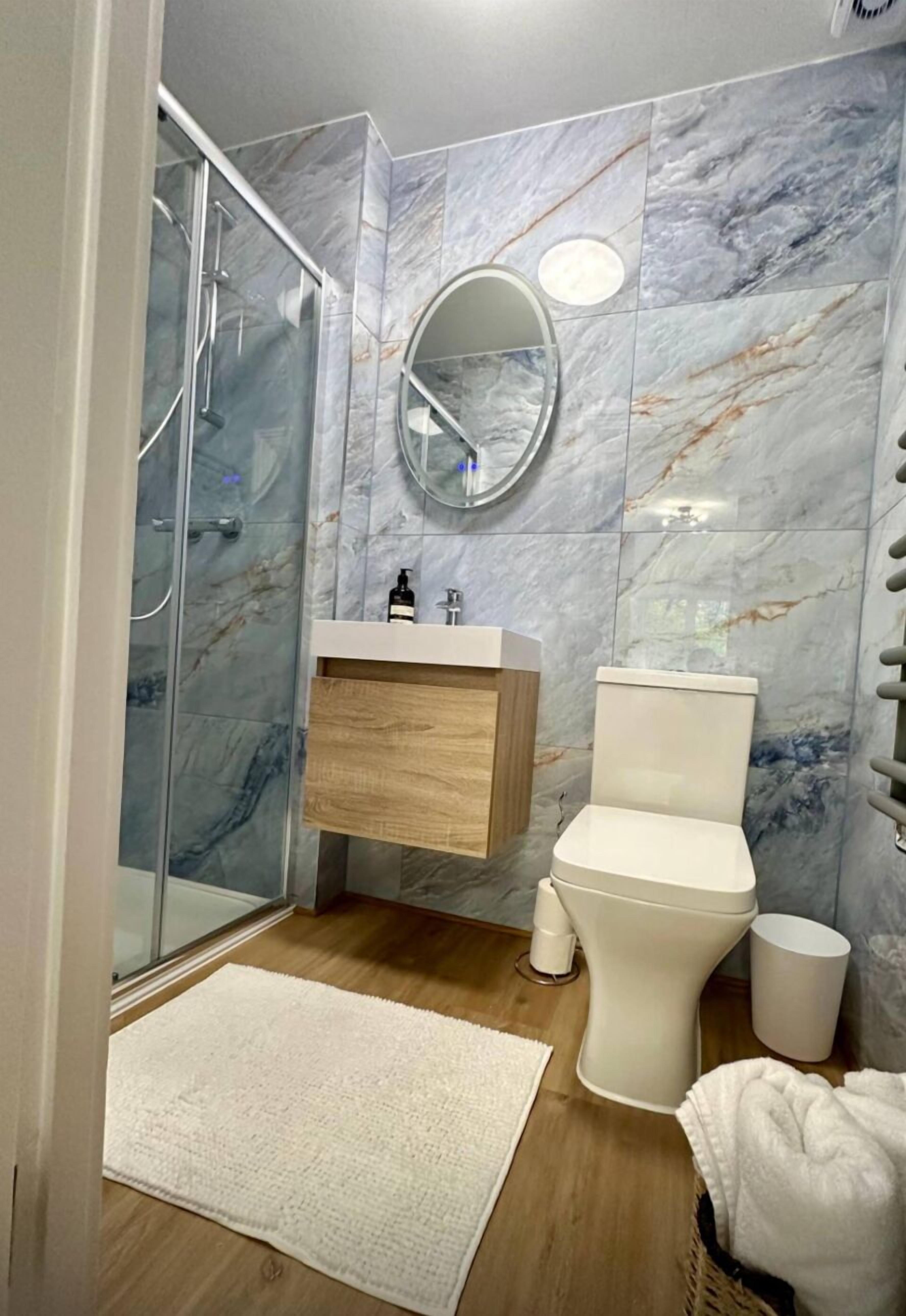 A bathroom with a glass shower enclosure, a wooden vanity with a mirror, a toilet, and a white rug on the floor.