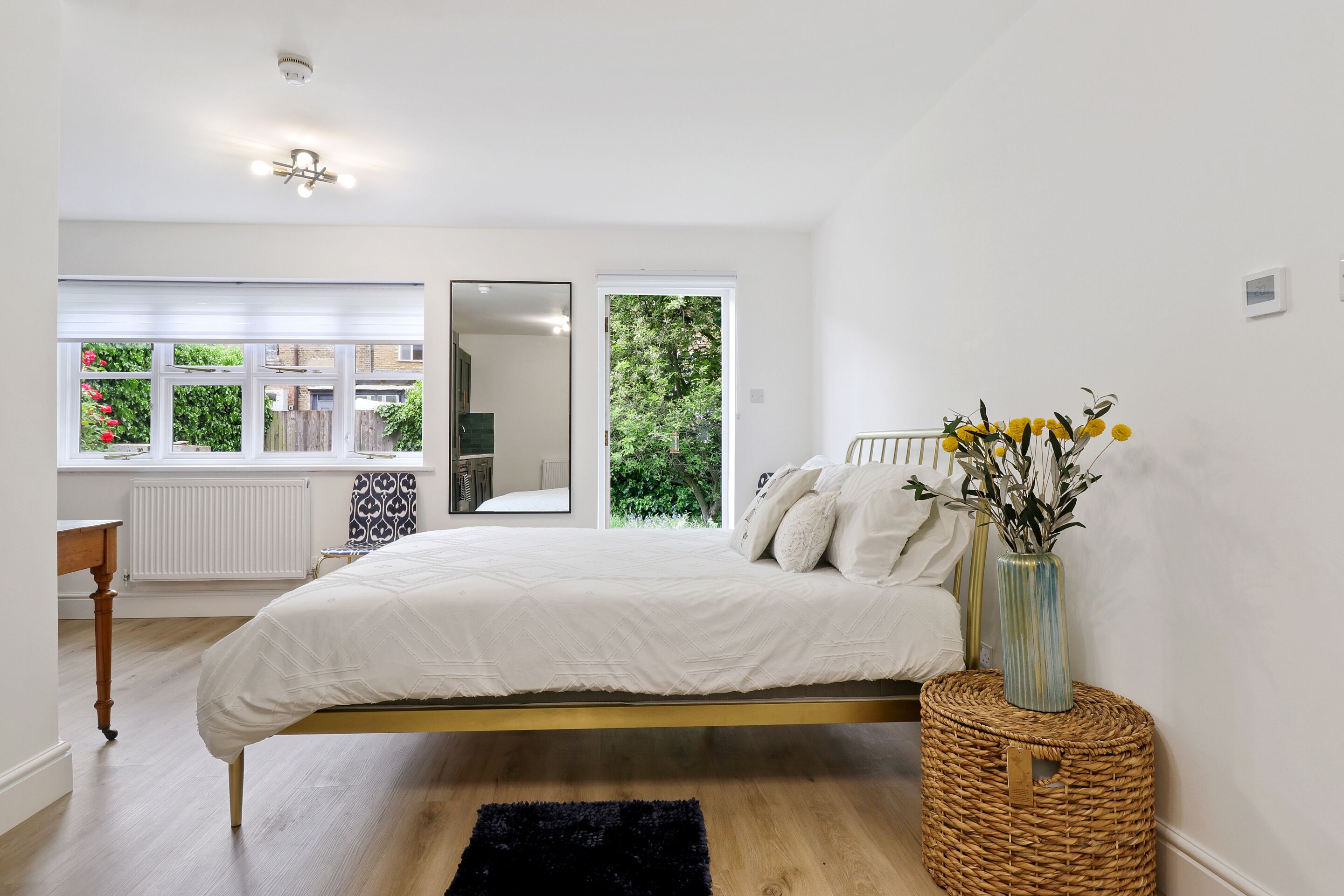 A bedroom with a bed, a window, a mirror, and a wicker basket with flowers.