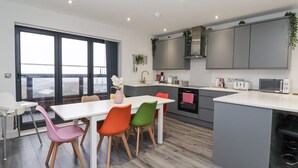 A modern kitchen with grey cabinets, a white countertop, and a built-in oven. There is a dining area with a white table and colorful chairs. - Seashell (Hornsea)