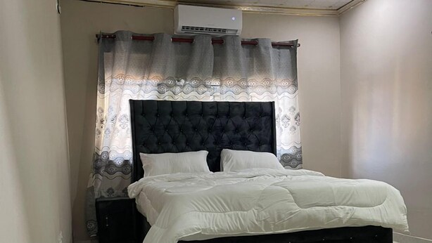 A bedroom with a bed, a nightstand, a wall-mounted air conditioner, and a patterned curtain. - ELITE HOME STAYS (Lusaka)