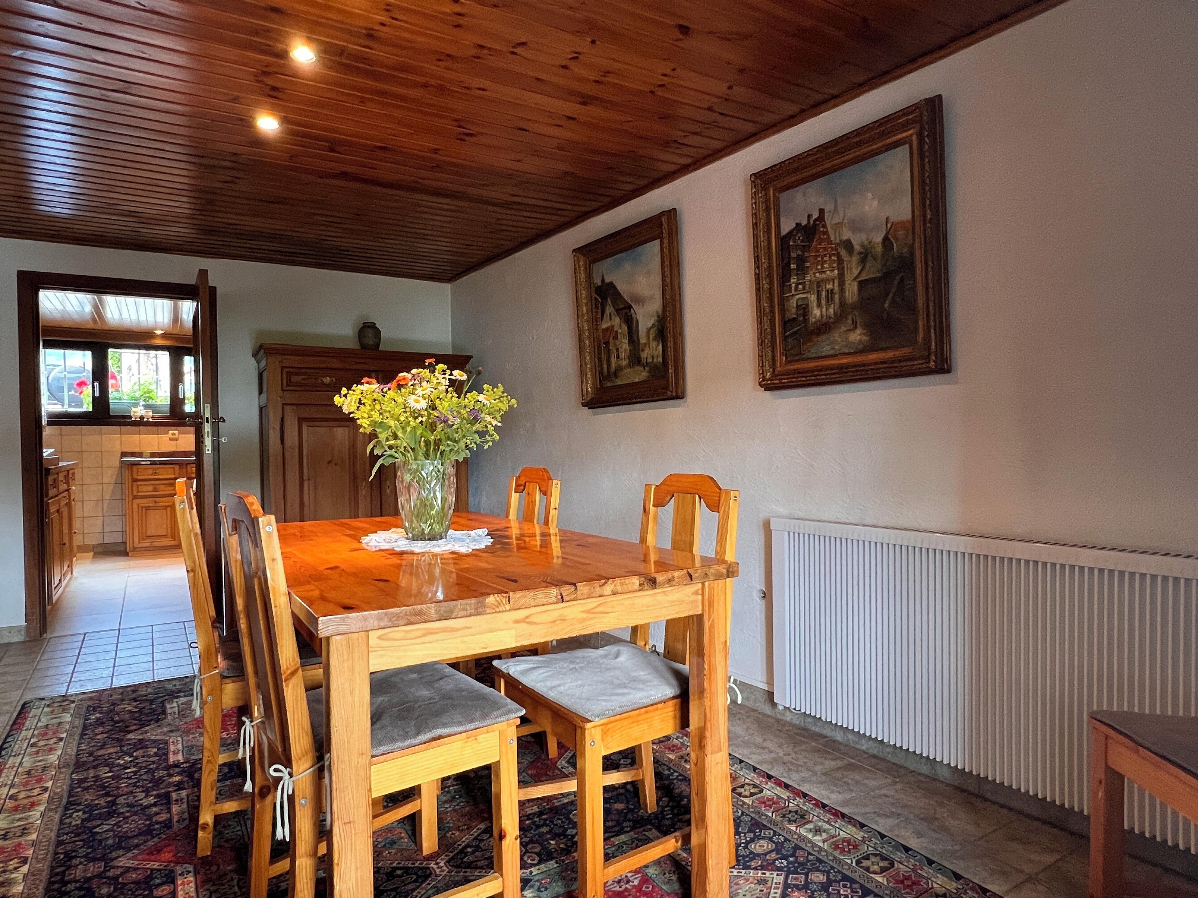 A dining room with a wooden table, chairs, a vase of flowers, and two framed paintings on the wall.