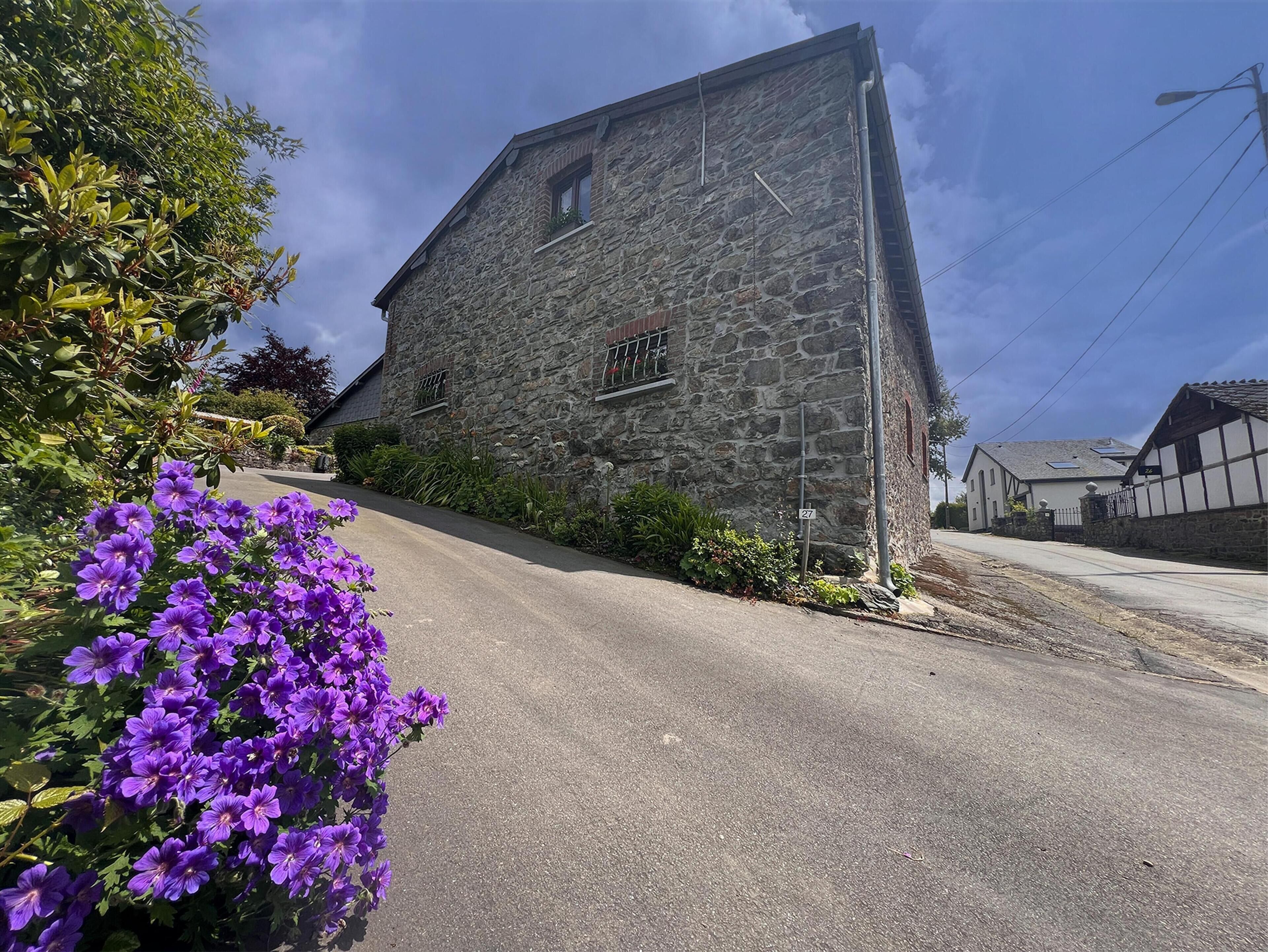 A stone building with a sloped roof, a narrow road in front, and purple flowers to the left.