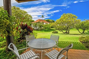 Une terrasse avec une table ronde en verre et des chaises en métal blanc, entourée d’une végétation luxuriante et offrant une vue sur une maison avec un toit rouge.