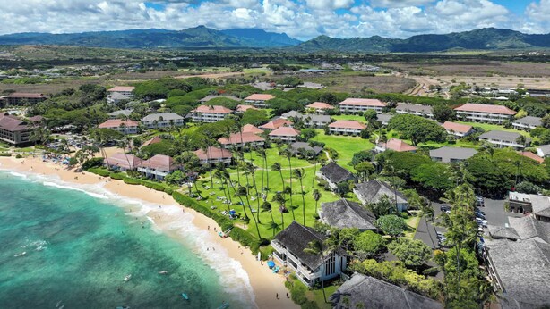 An aerial view of a coastal resort with multiple buildings, a beach, and mountains in the background. - Remodeled #56,  AC, grnd flr end unit, ideal location! (Koloa)