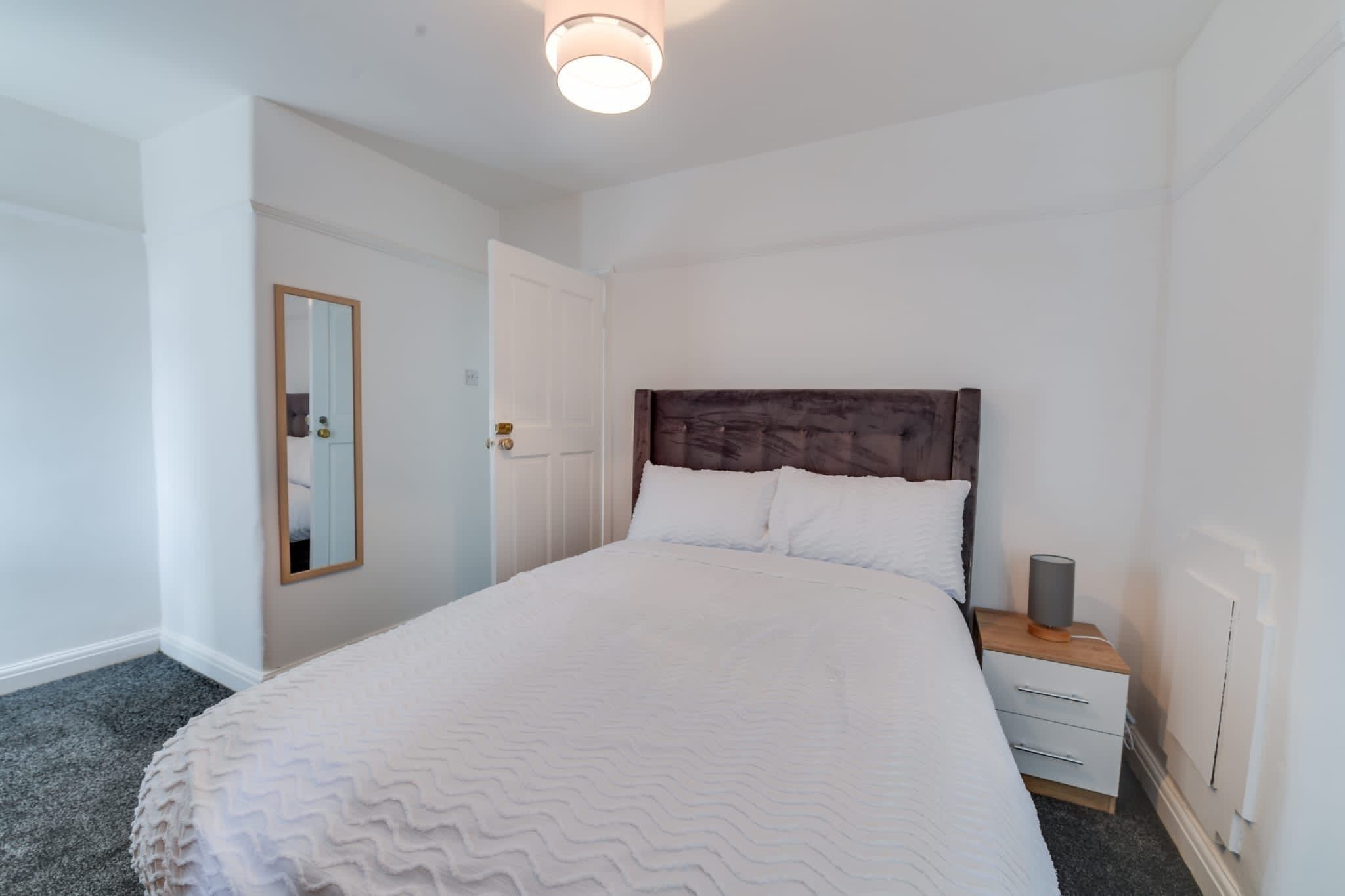 Ein Schlafzimmer mit einem großen Bett, einem Nachttisch mit Lampe, einem Spiegel und einer Tür.