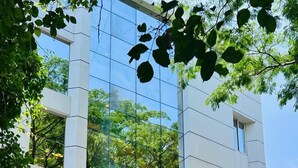 A modern hotel with a glass facade reflecting trees and buildings. There are two white cars parked in front. - Hotel Classio Kormangala (Bengaluru)