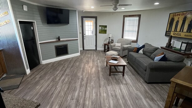 A spacious living area with a grey sofa, a wooden dining table, and a kitchen area with a refrigerator and a TV mounted on the wall.