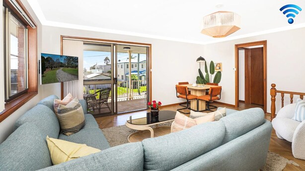A living room with a sofa, a coffee table, and a dining area with chairs and a table. There is a sliding glass door leading to a balcony with a view of houses. - Sylvia​’​s at Currarong – peaceful retreat (Currarong)