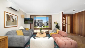 A living room with a grey sofa, a pink sectional, a coffee table with fruit, and a TV on the wall. - Sylvia​’​s at Currarong – peaceful retreat (Currarong)