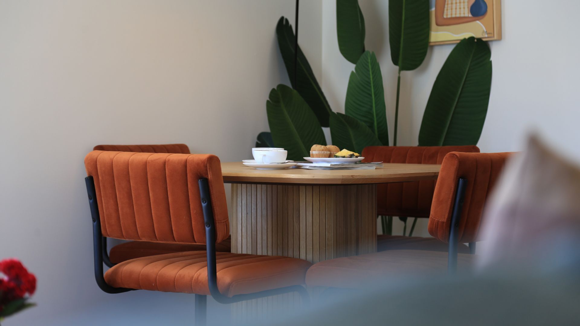 Una mesa redonda con cuatro sillas tapizadas en naranja, una taza blanca, un plato con comida y un muffin.