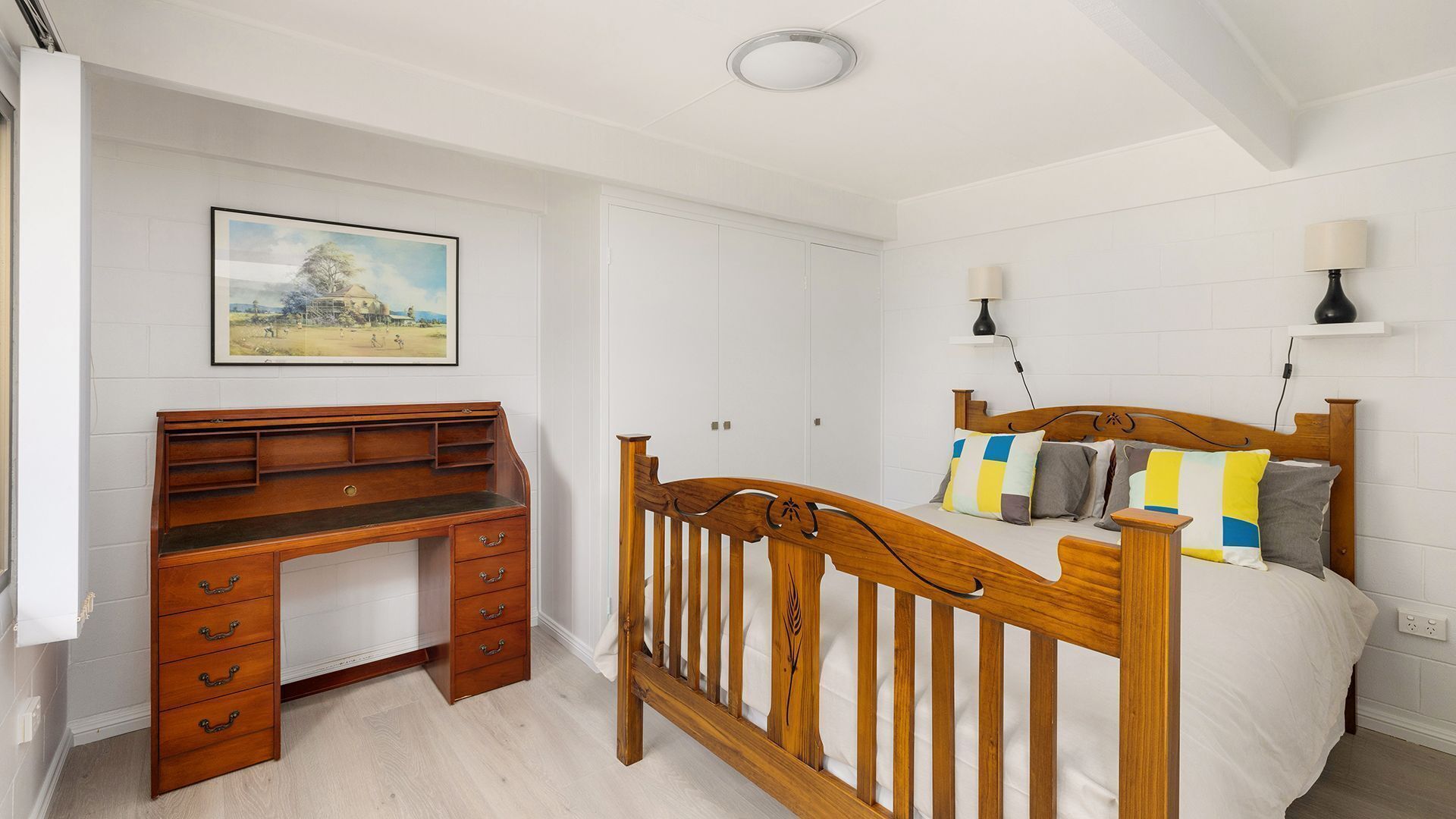 Une chambre avec un bureau en bois, un lit agrémenté de coussins colorés, une table de chevet et une lampe fixée au mur.