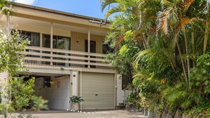 Un bâtiment à deux étages doté d’un balcon, d’un garage et d’une végétation tropicale luxuriante.