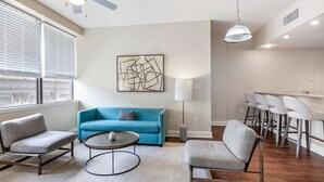 Ein modernes Wohnzimmer mit einem blauen Sofa, grauen Sessel und einem Couchtisch, sowie einem Essbereich mit Barhocker.
