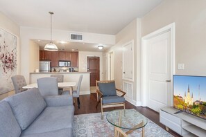 A modern living room with a grey sofa, a glass coffee table, and a TV displaying a cityscape. - NOLA Hideaway: Modern 1BR Condo with BBQ Patio (New Orleans)