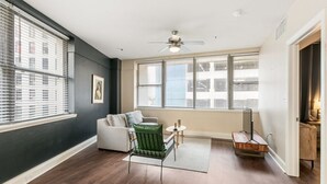 A room with a dark wall, a ceiling fan, a window with blinds, a sofa, a green chair, a small table with a lamp, and a wooden side table.