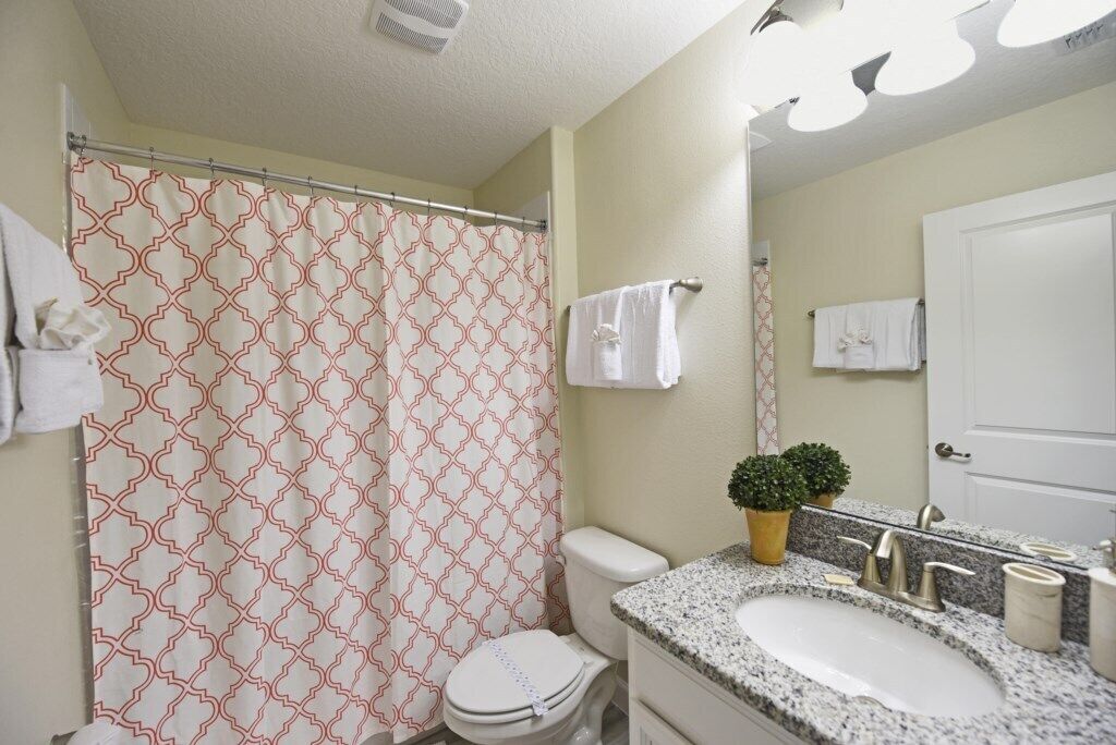Un baño con cortina de ducha estampada, inodoro, lavabo con encimera de granito, una planta pequeña en maceta y un espejo.