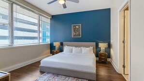 Une chambre à coucher avec un grand lit, deux tables de chevet avec des lampes, un ventilateur de plafond, une fenêtre avec des stores et une porte.
