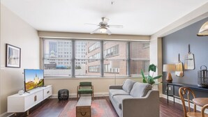 Ein modernes Wohnzimmer mit einer Couch, einem Couchtisch, einem Fernseher und einem großen Fenster mit Jalousien.