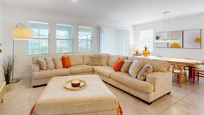 A modern living room with a beige sectional sofa, a matching ottoman, and a dining area with a table and chairs. - Beautiful 5 Bed Pool Home with Game Room (Davenport)