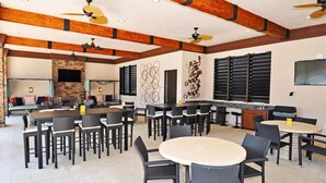 A spacious outdoor dining area with multiple tables and chairs, a stone fireplace, and a bar area with a television. - Beautiful 5 Bed Pool Home with Game Room (Davenport)
