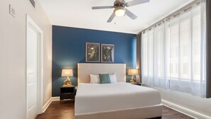 A bedroom with a large bed, two bedside tables with lamps, a ceiling fan, and a window with sheer curtains. - French Quarter Gem: Stylish 1BR Condo with Pool Oasis (New Orleans)