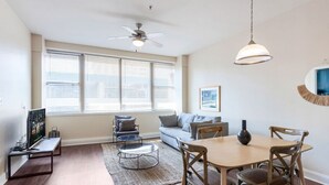 Un salon moderne avec un canapé, une table basse et une table à manger. On y trouve un ventilateur de plafond, de grandes fenêtres équipées de stores et un tableau encadré accroché au mur.