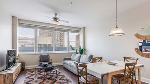 A modern living room with a sofa, dining table, and chairs. There is a ceiling fan, a TV, and a window with blinds.