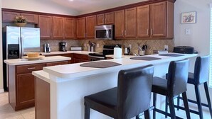 A modern kitchen with wooden cabinets, a stainless steel refrigerator, a microwave, and a coffee maker. There are two bar stools at the island. - Spacious Family Pool Home with Game Room (Davenport)