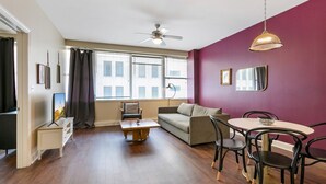 A modern living room with a sofa, coffee table, and dining area with chairs. There is a ceiling fan, large windows with blinds, and a view of buildings outside. - Urban Oasis: Modern 1BR Condo with Rooftop Pool (New Orleans)