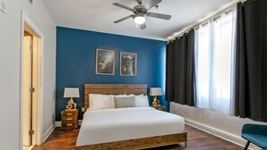 A bedroom with a wooden bed, two bedside tables with lamps, a ceiling fan, and a window with curtains. - Urban Oasis: Modern 1BR Condo with Rooftop Pool (New Orleans)