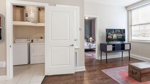 Un lavadero con dos lavadoras y una secadora, un televisor sobre un mueble, y una ventana con persianas.